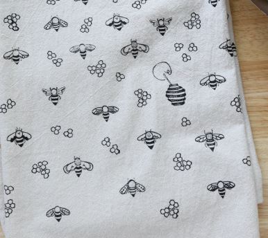 A close-up of a white fabric with a repeating black printed pattern of bees, honeycombs, and a beehive, laid flat on a light wood surface.