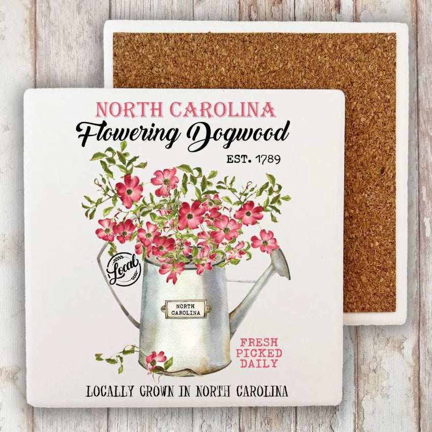 A square printed coaster or tile shown front and back on a whitewashed wood background. The front features a white base with a vintage-style illustration of pink flowers in a silver watering can, with text reading “NORTH CAROLINA Flowering Dogwood EST.