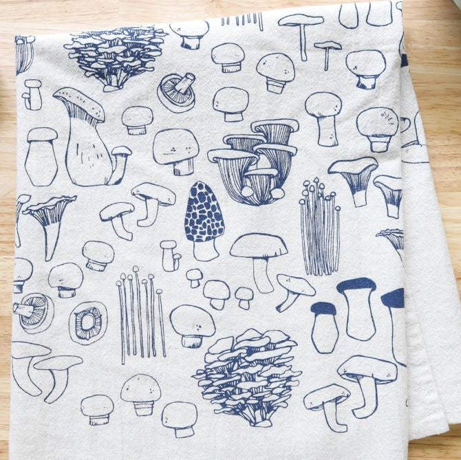 A white kitchen towel with a blue line-art print of various mushrooms, displayed on a light wood surface.