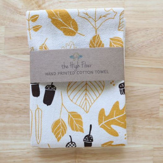A folded cotton towel with a white background and a gold, mustard yellow, and black leaf and acorn pattern, wrapped with a brown paper band that reads 'the High Fiber HAND PRINTED COTTON TOWEL' on a light wood background.