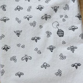 A close-up of a white fabric with a repeating black printed pattern of bees, honeycombs, and a beehive, laid flat on a light wood surface.