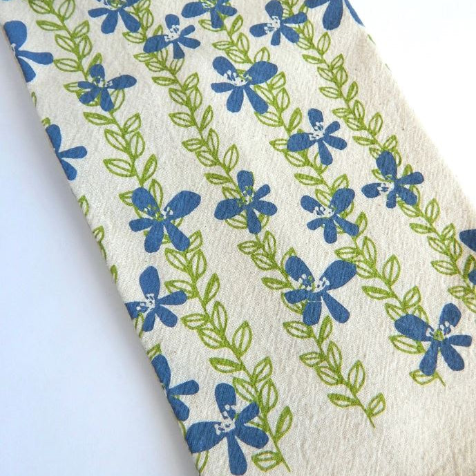 A close-up of a cream-colored fabric with a repeating pattern of blue flowers and green leaves, laid diagonally on a white background.