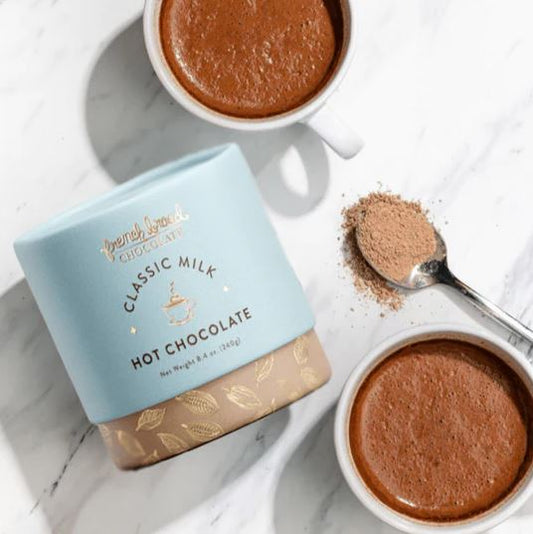 Two cups of hot chocolate with a box of Fair Trade Classic Milk Hot Chocolate on a marble surface.