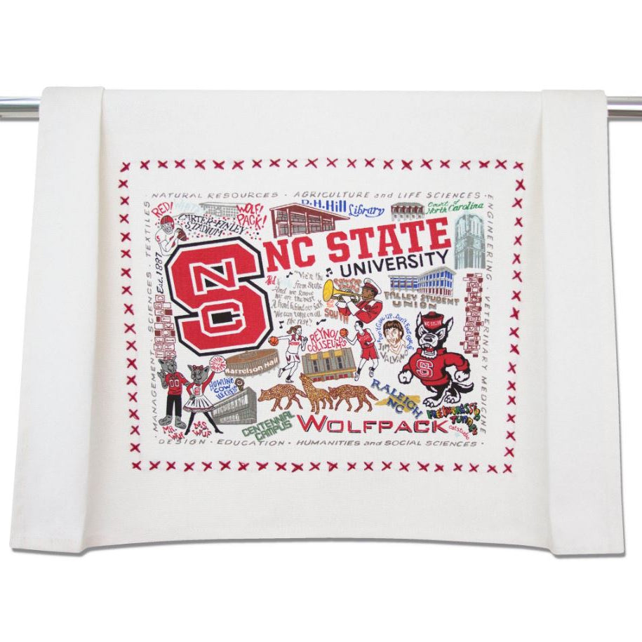 An off-white cotton tea towel featuring a detailed graphic print of NC State Wolfpack university themes, including the 'NC' logo, mascot, and campus landmarks, hanging from a metal rod.