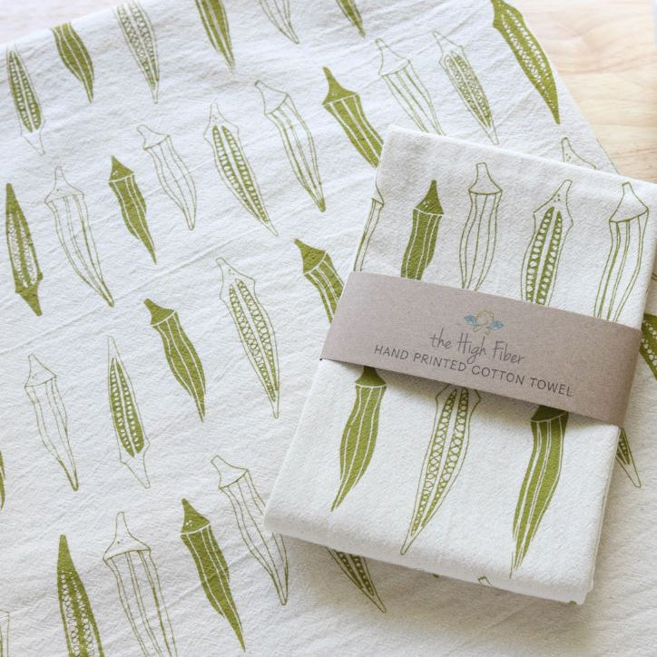 A folded white cotton towel with an all-over olive green and light gray botanical leaf pattern, wrapped with a brown paper band reading 'The High Flower HAND PRINTED COTTON TOWEL'.
