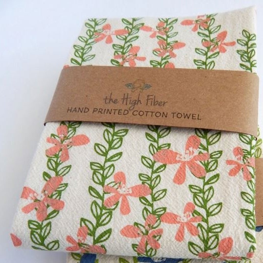 A folded cotton towel with a coral pink and green floral vine pattern, wrapped in a brown paper band labeled 'the High Fiber HAND PRINTED COTTON TOWEL'.